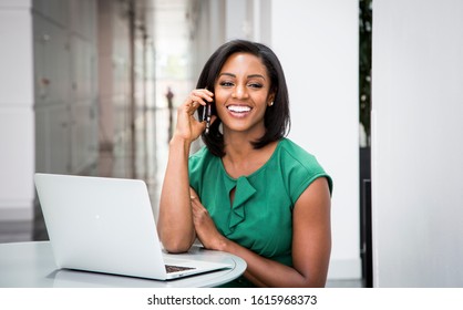 African American woman in green dress in talking on cellphone laughing with computer open in modern corporate building lobby 