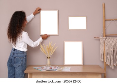 African American woman hanging empty frame on pale rose wall over table in room. Mockup for design