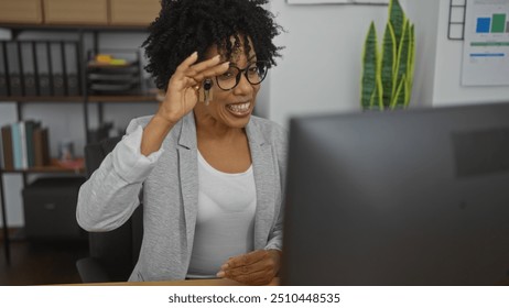 African american woman having a video call in an office, smiling behind a computer screen, wearing glasses and holding keys with a green plant and shelves in the background. - Powered by Shutterstock