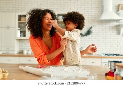 African American Woman With Her Little Daughter With Curly Fluffy Hair Having Fun And Cooking Pastries In The Kitchen. Mom And Daughter Cooking Together And Soiling Each Other With Flour.