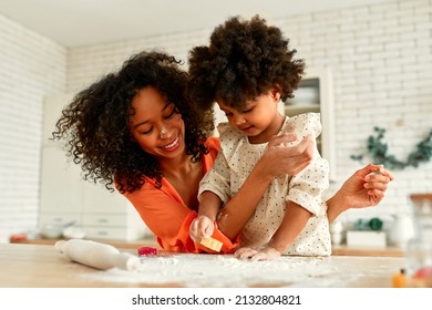 African American woman with her little daughter with curly fluffy hair having fun and cooking pastries in the kitchen. Mom and daughter cooking together. Child cutting out cookies with cookie cutters.