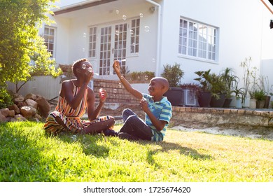 African American woman and her son, spending time together, sitting on a lawn, making soap bubbles. Social distancing and self isolation in quarantine lockdown.