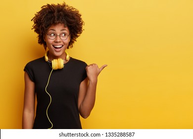 African American Woman In High Spirit Points Away With Thumb, Feels Entertained And Satisfied, Listens Radio Broadcasting With Modern Headphones Connected To Telephone, Checks Sound, Shows Free Space