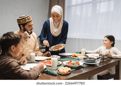 African american woman in hijab serving food on plate near family and ramadan dinner - Shutterstock ID 2266497783