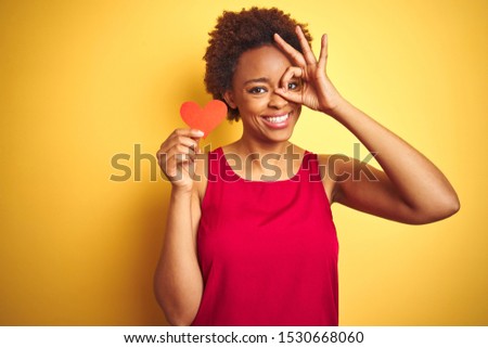 African american woman holding romantic paper hearts over yellow isolated background with happy face smiling doing ok sign with hand on eye looking through fingers