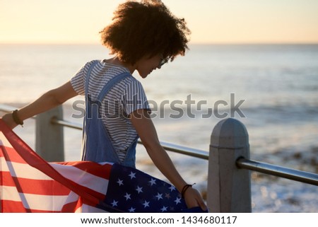 African american woman holding USA flag by the ocean, celebrating independence day