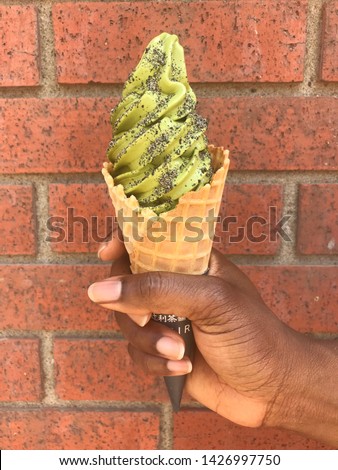African American woman holding a waffle cone with green ice cream and ground sesame against a brick building 