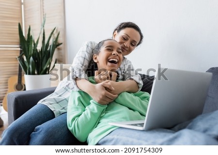 african american woman hugging laughing daughter while watching film on laptop