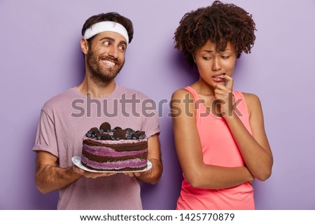 African American woman keeps finger on mouth, feels temptation while looks at delicious blueberry cake, tries not to eat unhealthy food, being on diet and goes in for sport. Mmm, how tasty it looks