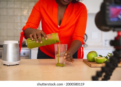 African American Woman In Kitchen Pouring Health Drink, Making Vlog Using Laptop And Camera. Online Cookery Vlogger At Home.