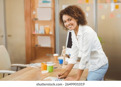 African American woman leaning over desk reviewing swatches with brush and paint jars in studio. Creative, artistic, modern, colorful, inspirational, workspace, design