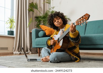 African American woman learning guitar watching tutorial on laptop at home. Black girl playing guitar singing song learning online music lessons. Artistic woman playing acoustic guitar learning chords - Powered by Shutterstock