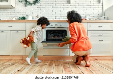 An African American Woman With A Little Daughter With Curly Fluffy Hair Is Taking Out A Baking Sheet With Baked Muffins From The Oven. Mom And Daughter Are Baking Cupcakes At Home In The Kitchen.