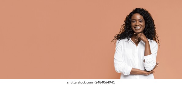 African American woman with long, curly black hair, wearing a white button-up shirt. She is smiling and has her arms crossed in front of her. The background is a peachy-orange color. - Powered by Shutterstock