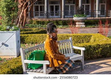 An African American woman with long sisterlocks wearing a brown coat and sunglasses sitting on a bench with lush green trees and plants at Atlanta Botanical Garden in Atlanta Georgia USA - Powered by Shutterstock