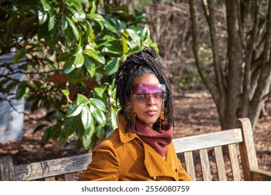 An African American woman with long sisterlocks wearing a brown coat and sunglasses sitting on a bench surrounded by lush green trees and plants at Atlanta Botanical Garden in Atlanta Georgia USA - Powered by Shutterstock