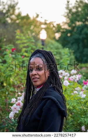an African American woman with long sisterlocks standing in the garden admiring the red roses in the garden surrounded by colorful flowers and lush green trees and plants at Atlanta Botanical Garden
