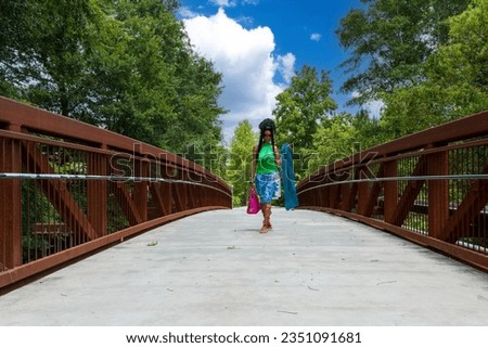 An African American woman with long sisterlocks wearing a green top, blue skirt carrying a pink purse on a bridge with lush green trees at the University of Georgia in Athens Georgia USA