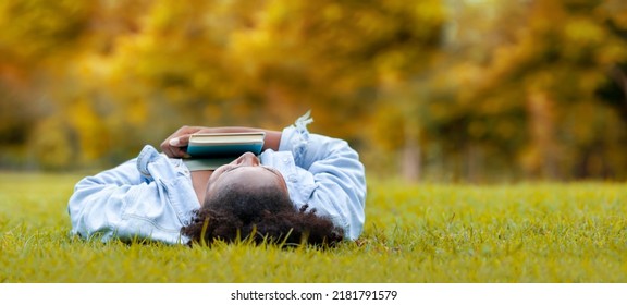 African American Woman Is Lying Down In The Grass Lawn Inside The Public Park Holding Book In Her Hand During Autumn For Fall Season And And Education Concept