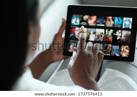 African American Woman Lying Down On Sofa At Home, Choosing Movie On Internet Streaming Service. Over The Shoulders.