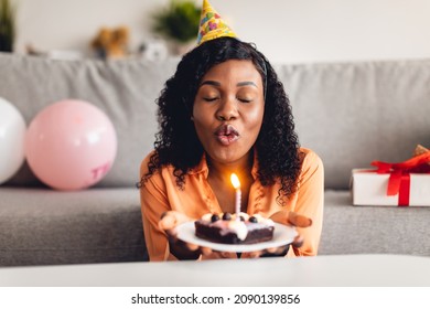 African American Woman Making Birthday Wish And Blowing Candle On Cake Wearing Festive Hat Sitting With Eyes Closed At Home. B-Day Holiday Celebration Concept. Front View