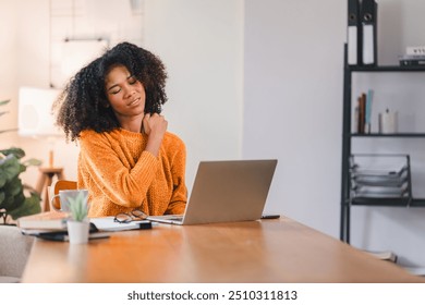 African American woman massaging her neck, indicating discomfort or strain while working on a laptop at home - Powered by Shutterstock