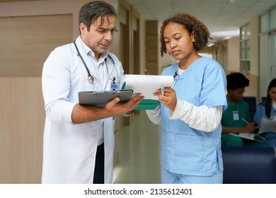 African American Woman Medical Student Is On The Job Learning With Professional Male Doctor At Medical School Hospital. Female Intern Doctor Holds Clipboard On The Job Training With Doctor Teacher.