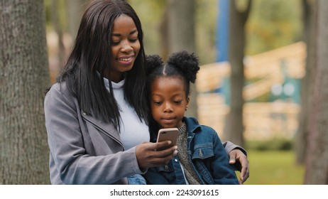 African American woman mother with child daughter little kid girl small baby schoolgirl sitting in park outdoors browsing mobile phone scrolling smartphone booking taxi order online with cellphone app - Powered by Shutterstock
