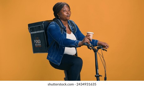 African american woman on bicycle enjoying cup of coffee before working her fast-paced delivery service job. Youthful female courier relaxing after on-demand package deliveries. - Powered by Shutterstock