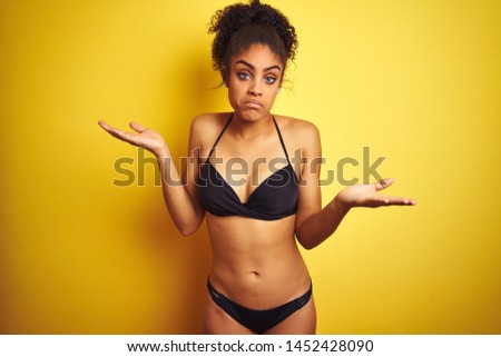 African american woman on vacation wearing bikini standing over isolated yellow background clueless and confused expression with arms and hands raised. Doubt concept.