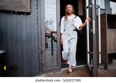 African American Woman In Overalls And Beret Walking Out The Door In Outdoor Terrace With Christmas Decorations.