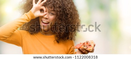 African american woman with a pizza slice with happy face smiling doing ok sign with hand on eye looking through fingers
