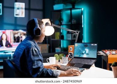 African American woman playing a competitive video game in her cozy living room using a wireless computer and wearing headphones. Young lady using keyboard, mouse and laptop for online gaming. - Powered by Shutterstock