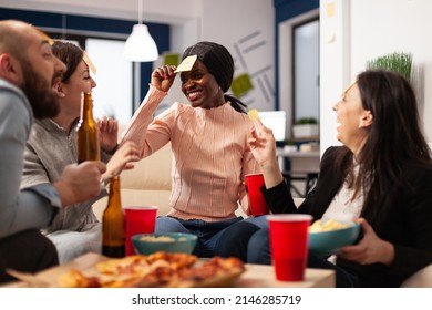 African American Woman Playing Guess Who Game With Coworkers, Celebrating With Drinks After Work. Office Colleagues Enjoying Charades Pantomime Play With Sticky Notes At Party.