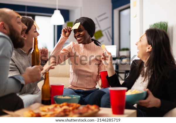 African\
american woman playing guess who game with coworkers, celebrating\
with drinks after work. Office colleagues enjoying charades\
pantomime play with sticky notes at\
party.