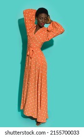 African American Woman Is Posing Barefoot With Hands Behind Head Wearing Orange Polka Dot Maxi Dress. Full Length Studio Shot On Turquoise Background.