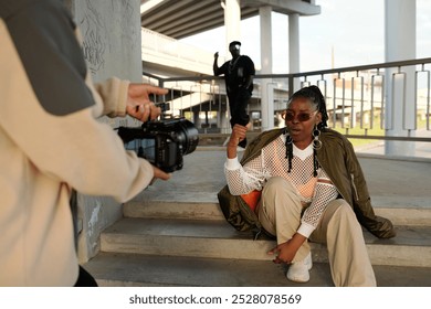 African American woman posing outdoors while photographer behind camera captures her posture under urban bridge, highlighting a casual and relaxed style - Powered by Shutterstock