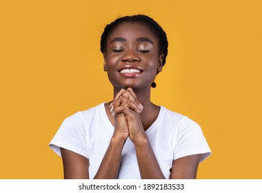 African American Woman Praying Holding Hands In Prayer Gesture Standing On Yellow Studio Background, Smiling With Eyes Closed. Black Female Prays. Hope Concept.