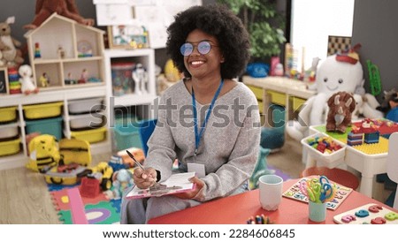 African american woman preschool teacher smiling confident writing on document at kindergarten