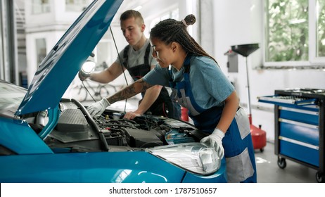 African american woman, professional female mechanic repairing car engine, tighten, screw with spanner while her colleague holding torch under car hood at service station. Side view. Web Banner - Powered by Shutterstock