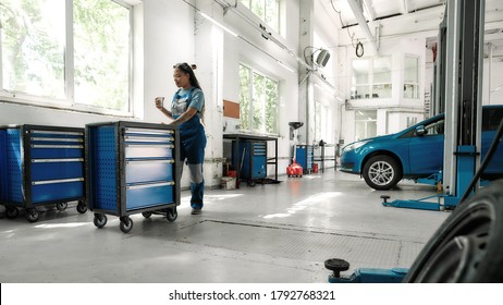 African american woman, professional female mechanic drinking coffee while pulling tool box cart in auto repair shop. Car service, maintenance and people concept. Selective focus. Web Banner - Powered by Shutterstock