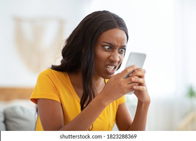 African American Woman Receiving Bad News, Angry At Her Smartphone At Home. Furious Black Lady With Mobile Phone Feeling Irritated Or Stressed Indoors