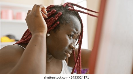 An african american woman with red braids in a room adjusting her hairstyle looking away from the camera - Powered by Shutterstock