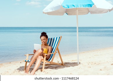 african american woman relaxing on deck chair and using digital tablet under beach umbrella in front of sea  - Powered by Shutterstock