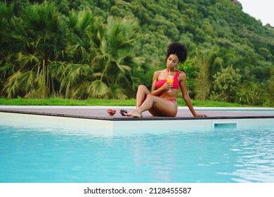 African American woman relaxing poolside drinking healthy smoothie and eating some fruit against a green hill in the summer - Powered by Shutterstock