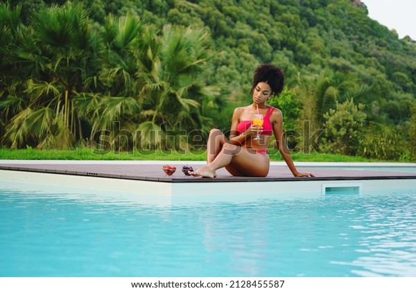 African American woman relaxing poolside drinking\
healthy smoothie and eating some fruit against a green hill in the\
summer