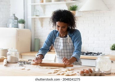 African American woman rolling out dough, cooking homemade cookies or pie at home, attractive young female housewife baker wearing apron baking pastry, using rolling pin, standing in cozy kitchen