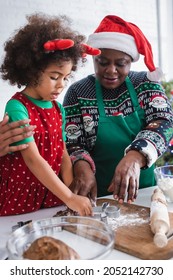 African American Woman In Santa Hat With Granddaughter In Reindeer Horns Headband Cooking In Kitchen