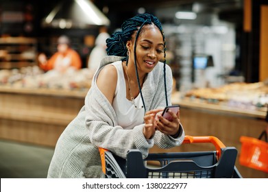 African American Woman With Shopping Cart Trolley In The Supermarket Store Look On Mobile Phone.