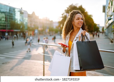 African American Woman Shopping With A Phone In European City Street At Sunset. Young Woman Standing Along The Road Holding Shopping Bags And Using Mobile Phone. Purchases, Black Friday, Discounts.